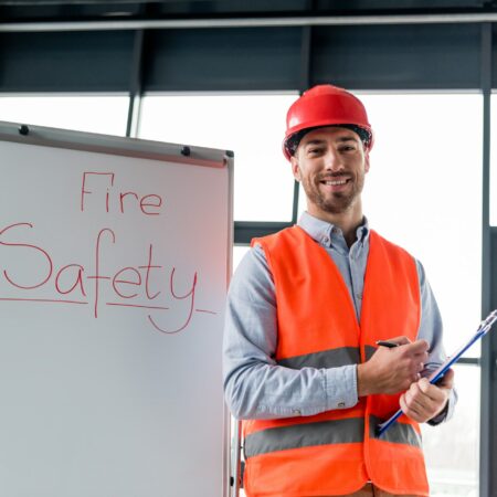 cheerful firefighter in helmet holding clipboard and pen while standing near white board with fire