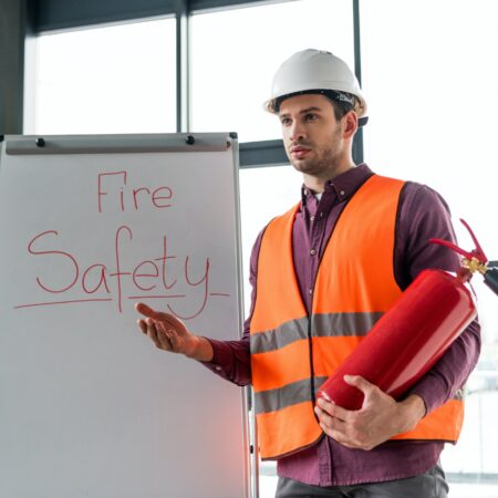 handsome fireman holding red extinguisher while standing near white board with fire safety lettering
